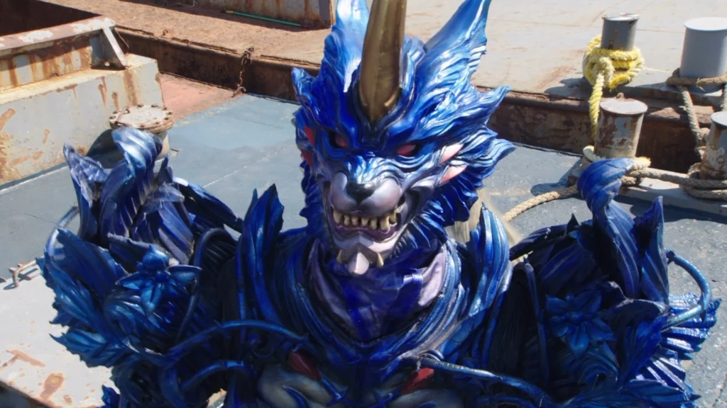 A scene from the Tokusatsu series featuring Kamen Rider Kiva. The character in focus is dressed in an intricate and formidable blue armor, resembling a mythical wolf. The helmet features sharp, menacing teeth, glowing red eyes, and a prominent golden horn on the forehead. The armor is detailed with wolf-like fur patterns and sharp edges, enhancing the fierce appearance. The background includes a weathered, industrial setting with rusted metal structures and thick ropes, adding a gritty contrast to the fantastical design of the Kamen Rider. The overall scene captures the intimidating and powerful presence of the character, ready for action in a rugged environment.