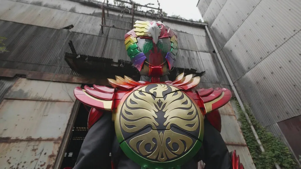 A scene from the Tokusatsu series featuring Kamen Rider OOO. The character is in full costume, wearing a striking, colorful armor with a prominent feline motif. The helmet is adorned with vibrant colors, including red, green, and yellow, and features a silver crest on top. The large, reflective eyes of the helmet add to the imposing appearance. The chest plate displays a large golden emblem of a mythical creature, possibly a phoenix, enhancing the regal and powerful look. The character stands in an industrial setting with rusted metal walls and a sense of decay, contrasting with the vibrant and polished armor. The overall impression is one of strength and heroism, typical of the Kamen Rider series.
