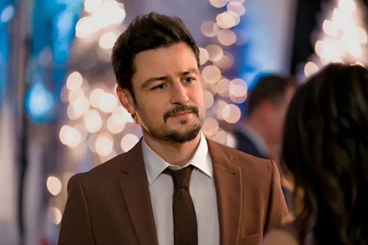 Tyler Hynes in a scene from "Never Been Chris'd," wearing a brown suit and tie, with a thoughtful expression on his face. He is standing in front of a festive, softly-lit background filled with twinkling lights, creating a warm and romantic atmosphere.