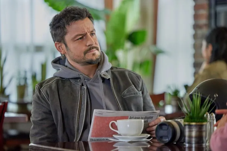 Tyler Hynes in a scene from "A Picture of Her," sitting in a cozy café. He is holding a newspaper with a curious expression, dressed in a grey hoodie layered under a leather jacket. On the table in front of him is a cup of coffee, a camera lens, and some potted plants, suggesting a relaxed yet intriguing atmosphere.