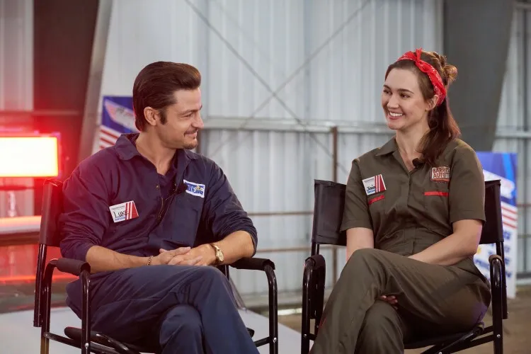 Tyler Hynes in a scene from "Shifting Gears," sitting alongside a female co-star in a garage setting. Both are dressed in mechanic jumpsuits; Tyler is wearing a navy blue jumpsuit, while the woman is in a green jumpsuit with a red bandana tied around her hair. They are seated in director-style chairs, smiling and engaged in conversation, suggesting a friendly and collaborative atmosphere.