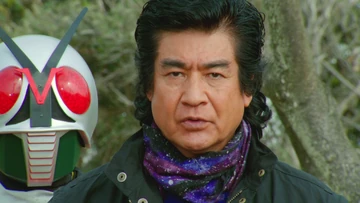 A scene from a Tokusatsu series featuring a middle-aged man with black hair and a serious expression, wearing a black jacket and a purple galaxy-patterned scarf. Behind him, partially visible, is a character in a white helmet with large red eyes, likely a Kamen Rider. The background consists of trees, suggesting an outdoor setting. The overall tone of the image is dramatic and intense, fitting the style of a Tokusatsu show.