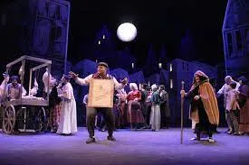 A theatrical production set under a full moon features a bustling town square scene, with performers dressed in period costumes engaging in lively activity. A man in the foreground commands attention, holding a prop or sign, while others around him add depth and drama to the scene, creating a vibrant and immersive stage atmosphere.