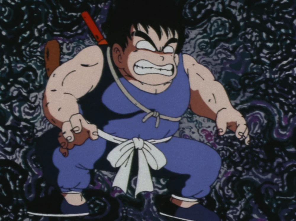 Goku, in his blue gi, crouches in an intense, angry stance, gripping his fists tightly. His expression is fierce and determined, set against a swirling, dark, and chaotic background.