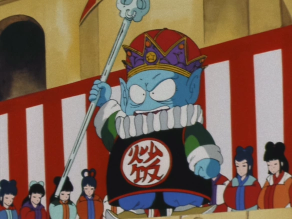 Emperor Pilaf stands proudly on a platform, wearing a crown and holding a staff, with a mischievous expression. He is surrounded by attendants dressed in traditional clothing, and the scene is framed by red and white striped banners.