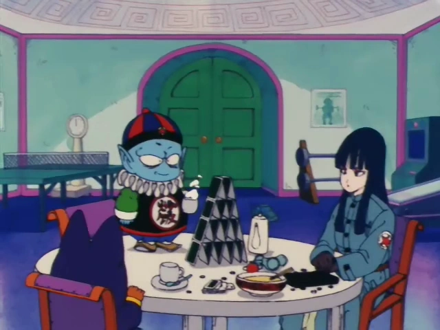 Emperor Pilaf smugly admires a card tower he's building on a table while Mai sits nearby with a bored expression. Shu is partially visible, seated on the opposite side. The room features a green double door, a clock, and a mix of furnishings in a whimsical setting.