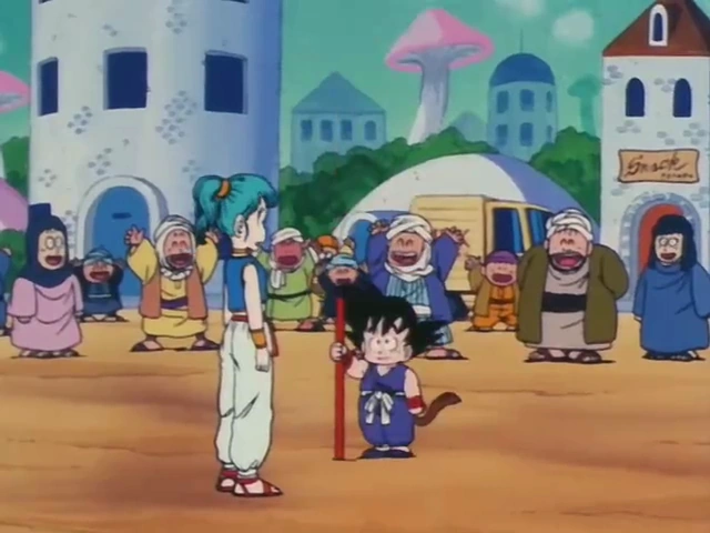 Bulma and Goku stand in the center of a lively town square, with Goku holding his Power Pole. The townspeople, dressed in colorful clothing, cheer and celebrate in the background, surrounded by domed buildings and mushroom-shaped trees under a green sky.
