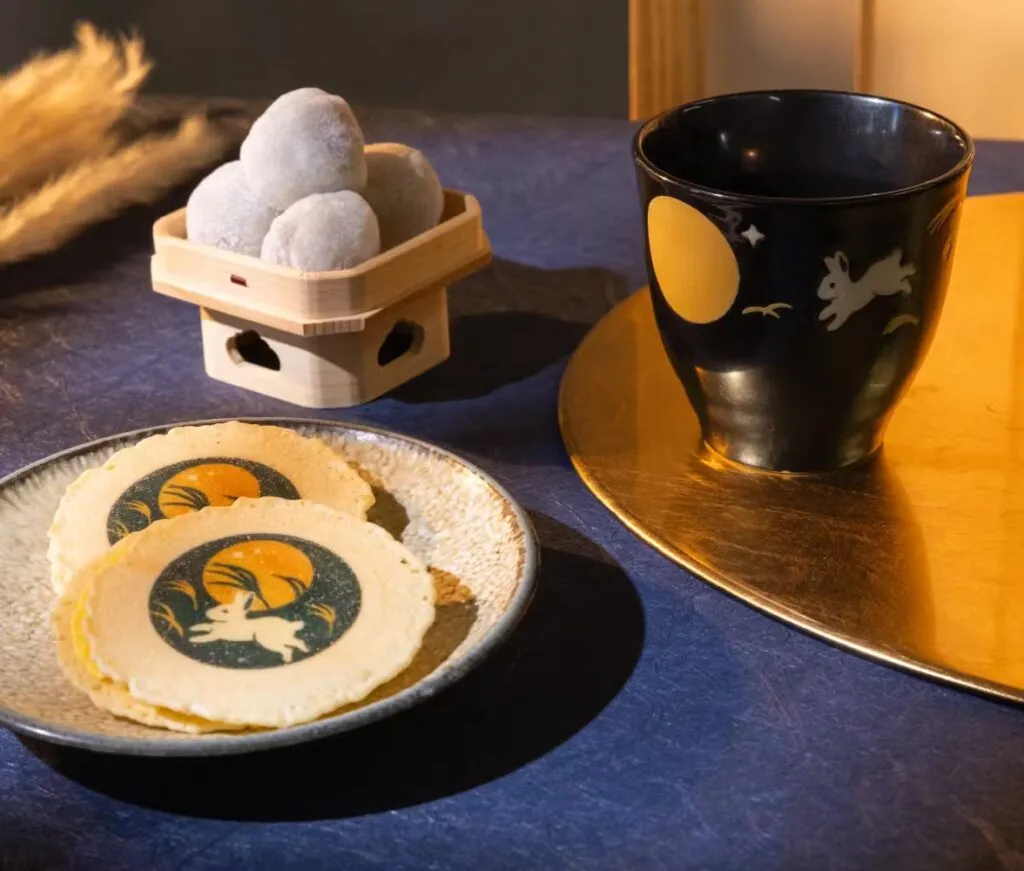 A black teacup decorated with a moon and rabbit design, a plate of rabbit-printed senbei rice crackers, and a wooden tray of round mochi displayed on a blue and gold table setting.