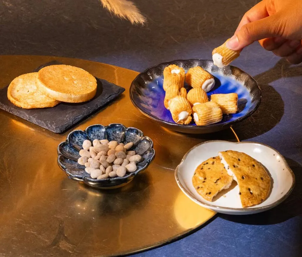 An assortment of Japanese snacks from the Sakuraco Autumn Moon Festival Box, including cream-filled crunchy rolls, sesame rice crackers, round senbei, and salted truffle-coated nuts displayed on ceramic plates.