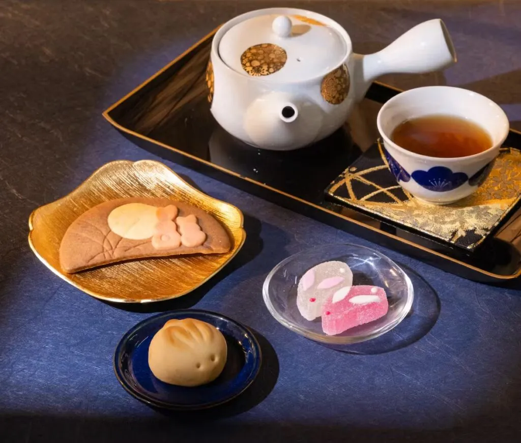 A Japanese tea set with a teapot, cup of tea, and seasonal tsukimi sweets including a rabbit-shaped manju, a moon and rabbit cookie, and rabbit-shaped jelly candies.