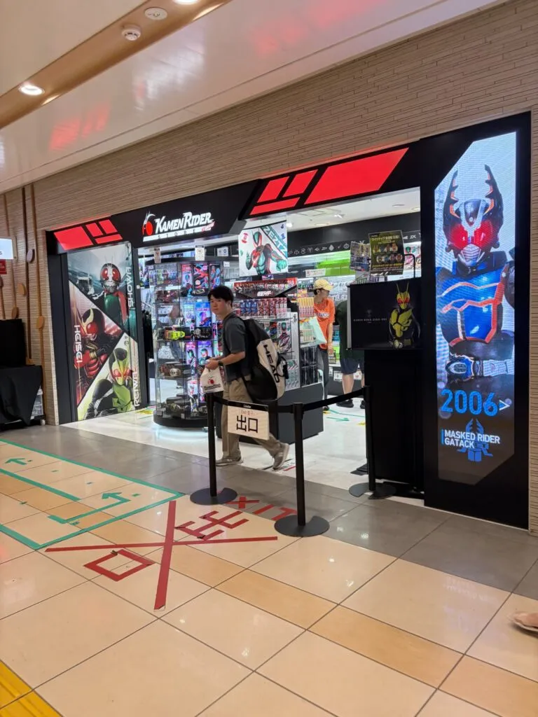 Entrance of the Kamen Rider Store Japan at Tokyo Station Ichibangai, featuring posters, LED displays, and Kamen Rider statues with fans browsing merchandise inside.