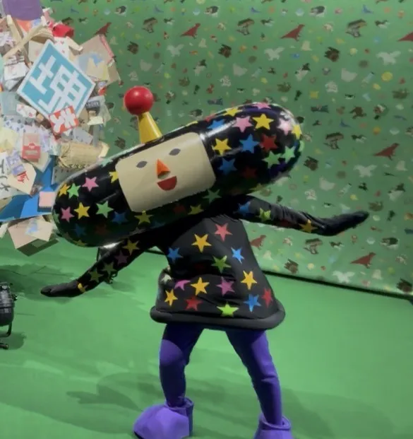 A colorful Katamari Damacy mascot dressed in a black outfit with multicolored stars poses playfully at Tokyo Game Show 2025 against a green background decorated with whimsical patterns and game art.