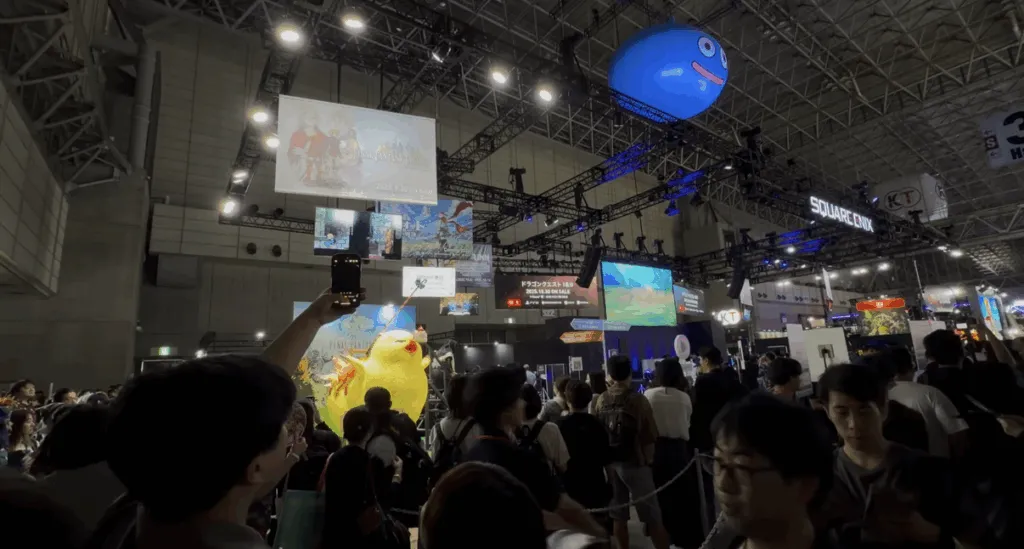 Crowds gather at the Dragon Quest booth during Tokyo Game Show 2025, featuring a giant inflatable blue Slime hanging above, large video screens, bright lights, and colorful displays across the bustling convention floor.