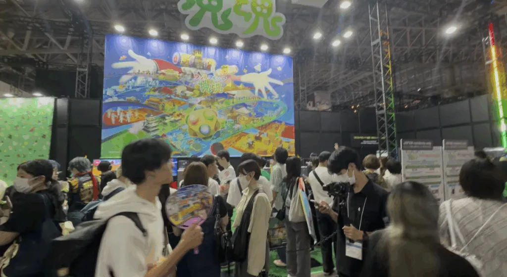 Attendees explore a colorful booth at Tokyo Game Show 2025 featuring a large vibrant display of a theme park-style game map, surrounded by bright lights, cheerful signage, and visitors holding fans and cameras.