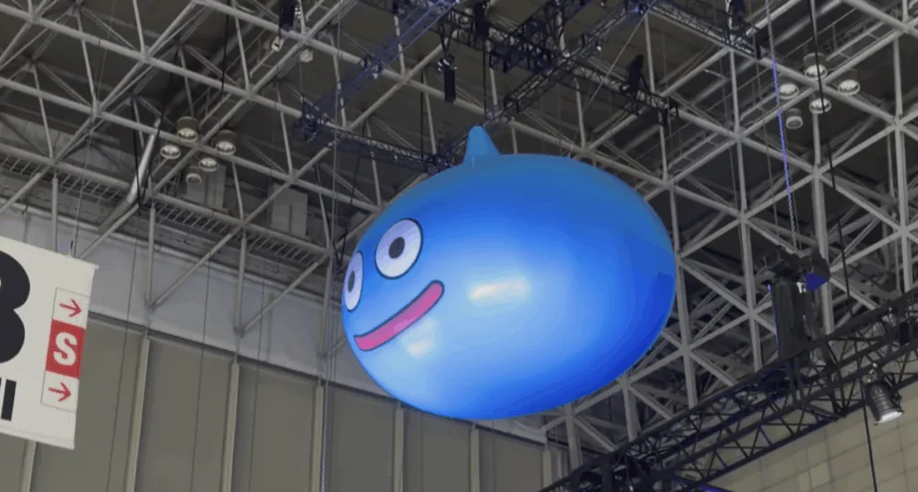 A giant inflatable blue Slime from Dragon Quest hangs from the ceiling at Tokyo Game Show 2025, smiling brightly under the metal beams and spotlights of the convention hall.