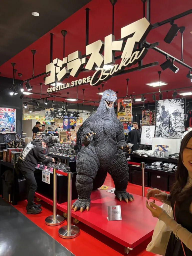 Life-sized Godzilla statue at the entrance of Godzilla Store Osaka inside Shinsaibashi PARCO, surrounded by merchandise and colorful posters.