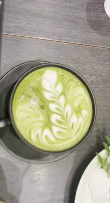 A beautifully crafted matcha latte with intricate leaf-style latte art served in a black cup on a dark wooden table. From Iyemon Salon Umeda.