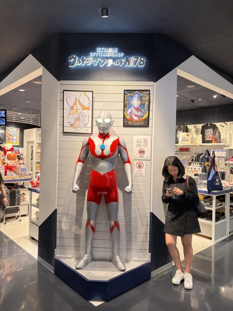 Life-size Ultraman statue at the entrance of Ultraman World M78 Shinsaibashi Store in Osaka, Japan, with a visitor standing nearby looking at their phone.