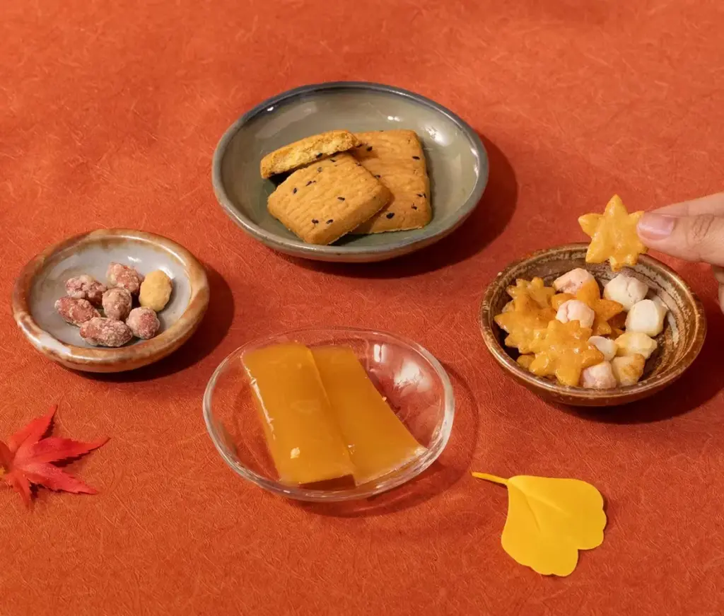 A colorful autumn-themed snack spread featuring Japanese treats like star-shaped rice crackers, sesame biscuits, candied nuts, and amber jelly sweets inspired by seasonal harvest flavors.