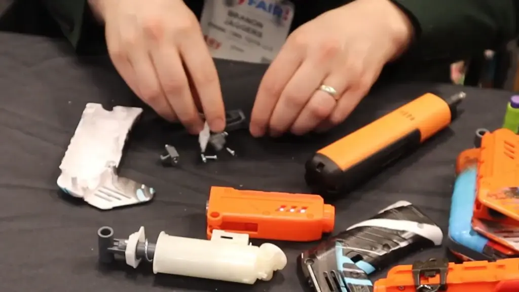 Close-up photo of a Dart Zone representative assembling a blaster at Toy Fair NY 2025, showing internal components, shell pieces, and a screwdriver on a black table.