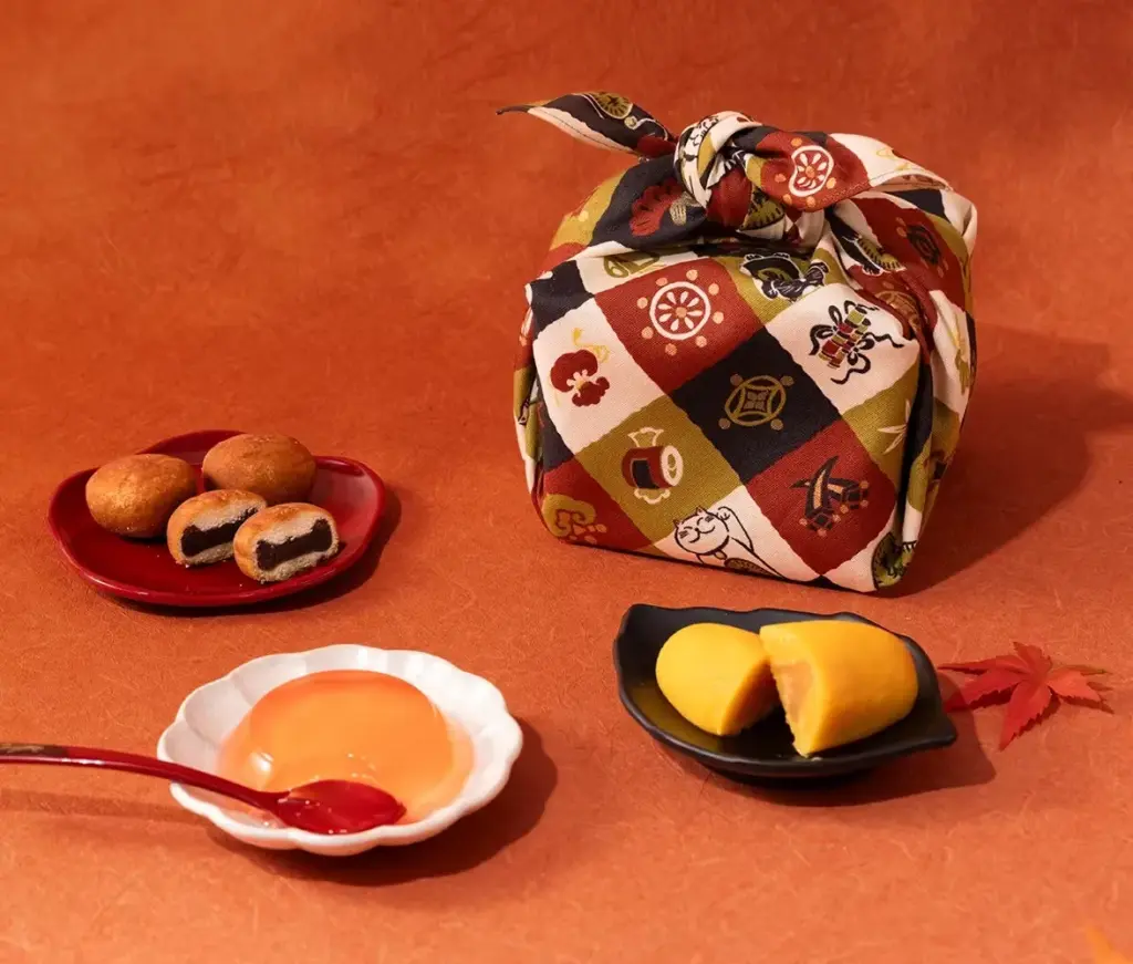 A cozy autumn dessert spread featuring Japanese sweets like Strawberry Anko Donuts, Apple Sweet Potato Cake, and a glossy jelly treat beside a decorative furoshiki-wrapped gift, all set against a warm orange background.