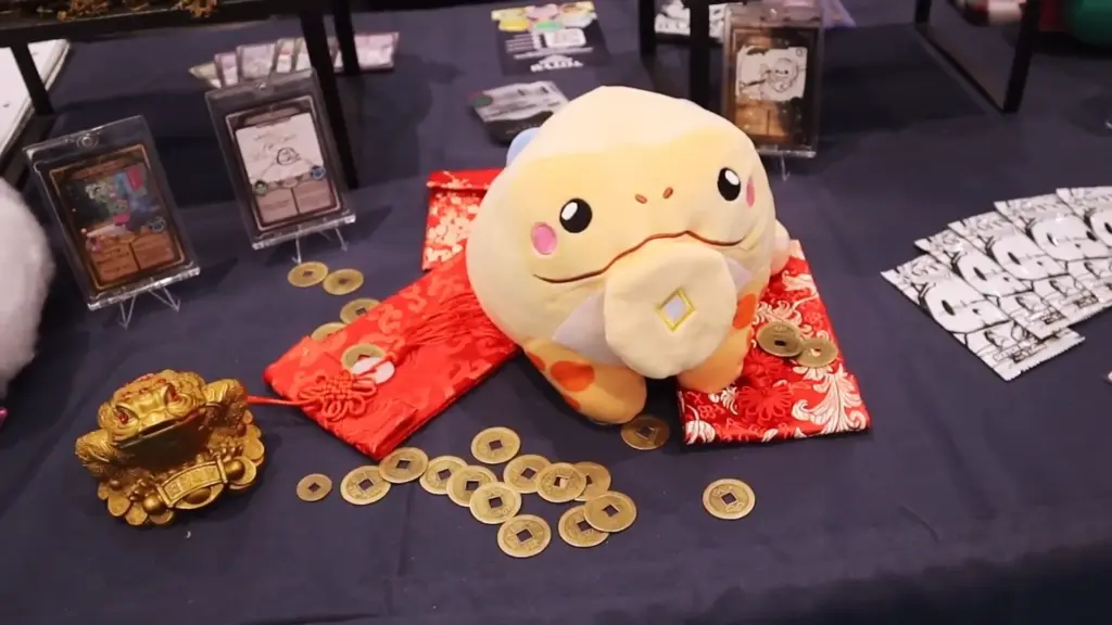Lucky the three-legged money toad plush from Totym Plush displayed at Toy Fair NY 2025, sitting on a red silk cloth surrounded by gold coins and collectible cards.