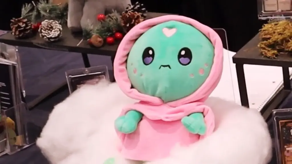 Luna the Dream Walker plush from Totym Plush at Toy Fair NY 2025, a green plush wearing a pink robe and hood, displayed on a bed of soft white cloud-like cotton.