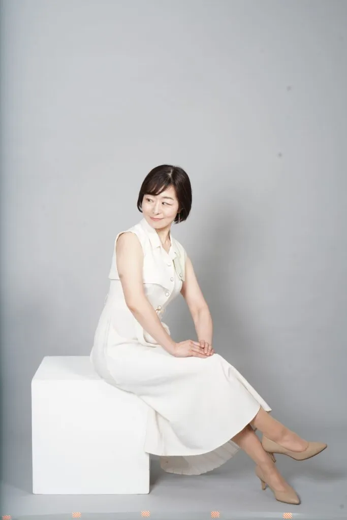 Mayuko Itagaki posing in a white outfit against a light background, smiling softly for a professional photoshoot.
