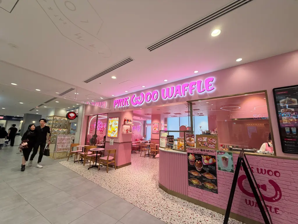 Exterior view of PINK WOO WAFFLE café in Japan with bright pink signage, cozy seating area, and cute pastel interior design.