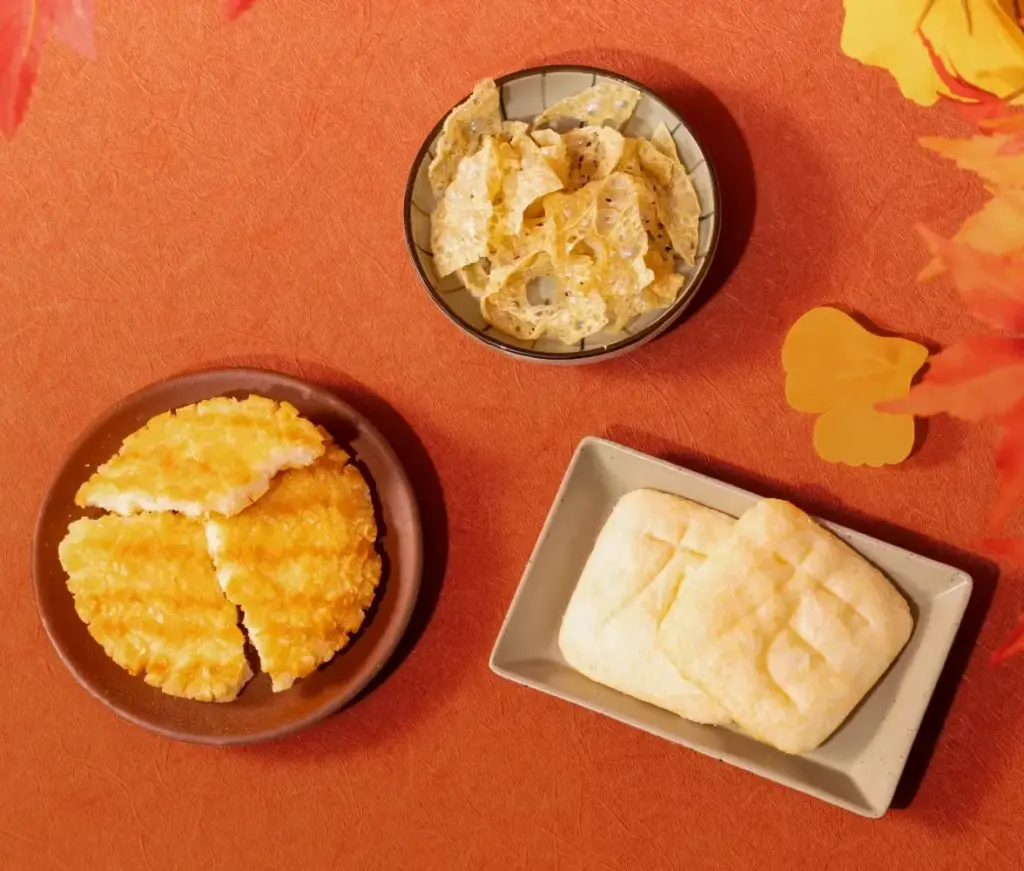 A display of savory Japanese snacks from Nikko including Nikko Yuba Chips, Nikko Jingoro Senbei, and crispy rice crackers arranged on autumn-themed dishes.