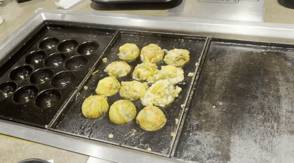 A close-up of takoyaki cooking on a hot griddle at Takonotetsu Tsunoda in Osaka, Japan, showing half-formed octopus balls being cooked in the pan.