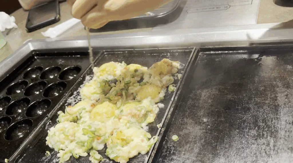 A hand turning takoyaki batter on a hot grill at Takonotetsu Tsunoda in Osaka, showing the interactive DIY cooking experience.