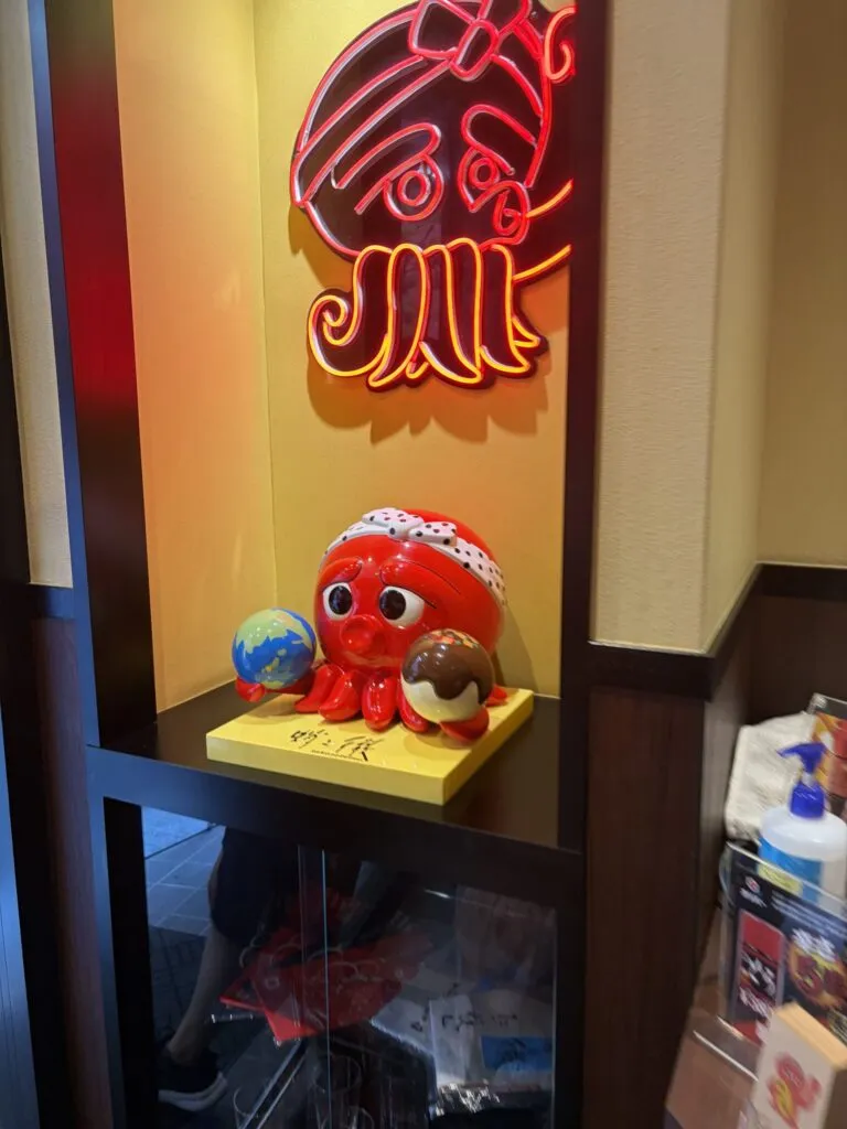 A cute red octopus mascot holding takoyaki and a globe, displayed inside Takonotetsu Tsunoda in Osaka, Japan, with a glowing neon sign above it.