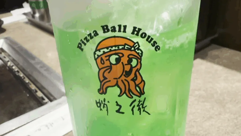 A glass of bright green melon soda with the Pizza Ball House logo, served at Takonotetsu Tsunoda in Osaka.