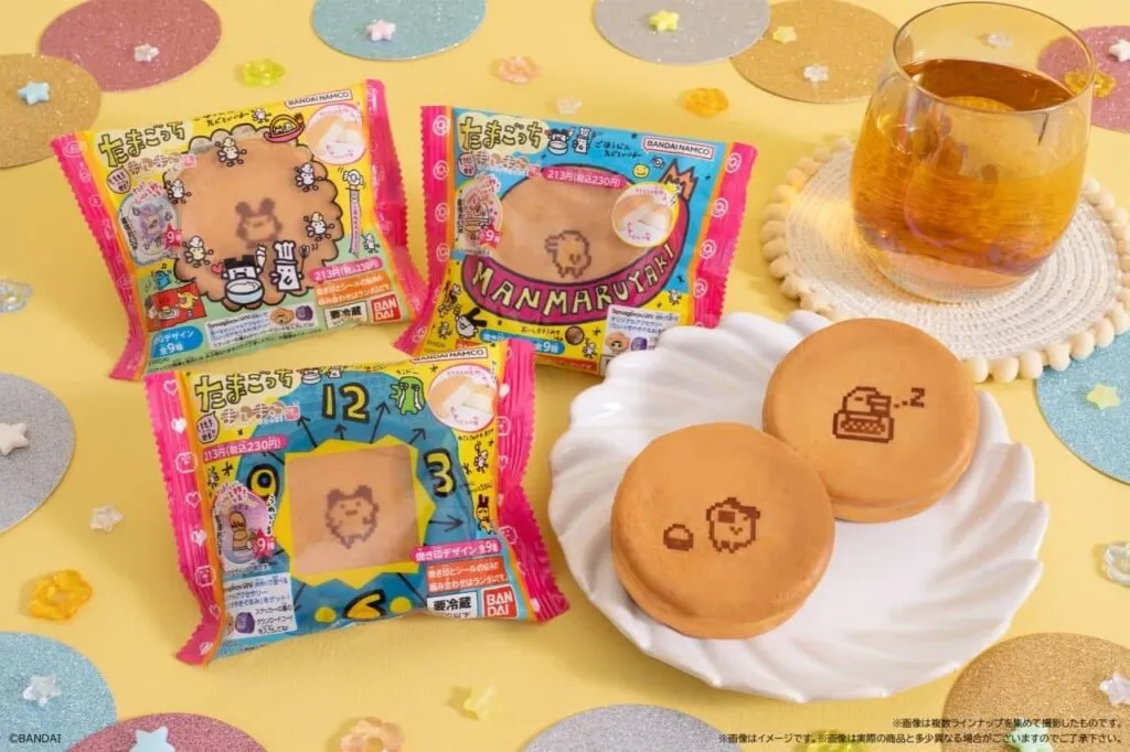 A colorful display of Tamagotchi Manmaruyaki snacks from Bandai Namco, featuring cute pixel Tamagotchi character designs on fluffy pancake treats, with a glass of tea beside them on a yellow background.