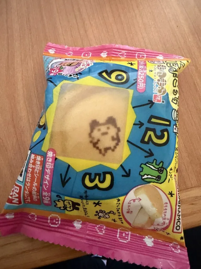 A packaged Tamagotchi Manmaruyaki snack with colorful Bandai Namco branding and Tamagotchi characters on the wrapper.