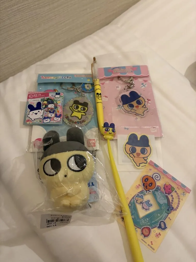 A collection of Tamagotchi merchandise including plush toys, keychains, a pen, and stickers featuring classic characters displayed on a hotel bed.