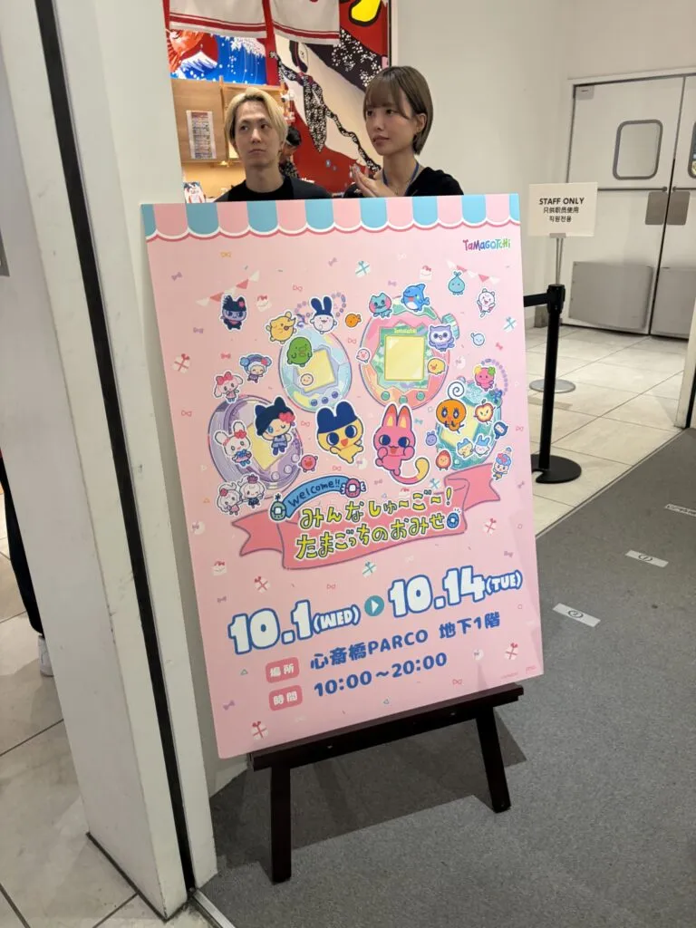 Entrance of the Tamagotchi pop-up store featuring a large pink signboard with character artwork and event dates, staffed by friendly employees.