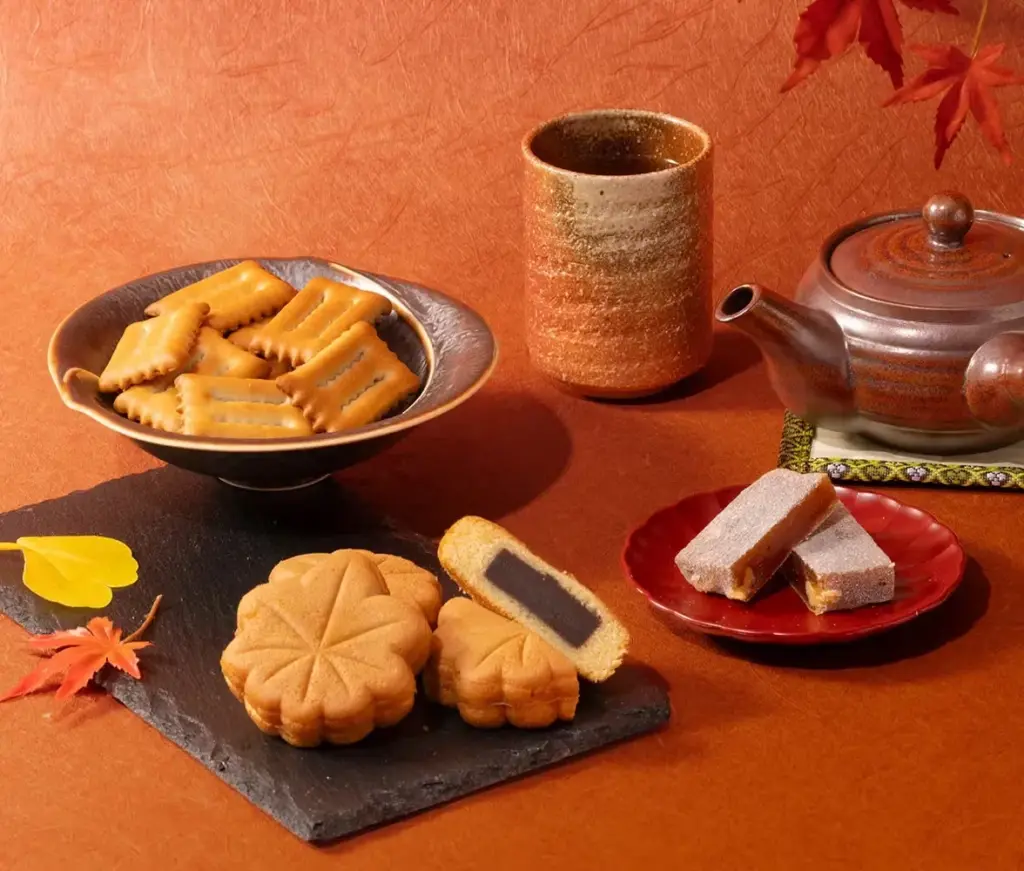 A cozy autumn tea setup featuring a warm cup of tea, a Japanese teapot, and an assortment of seasonal sweets including Momiji Manju, Walnut Mochi, and biscuits on a fall-themed table.