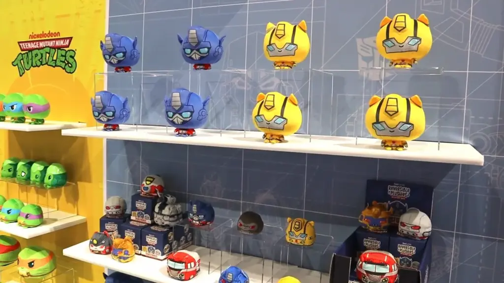 Shelves showcasing TeeTurtle reversible plushies inspired by Transformers characters including Optimus Prime and Bumblebee, displayed in blue and yellow on a branded wall at Toy Fair NY 2025.