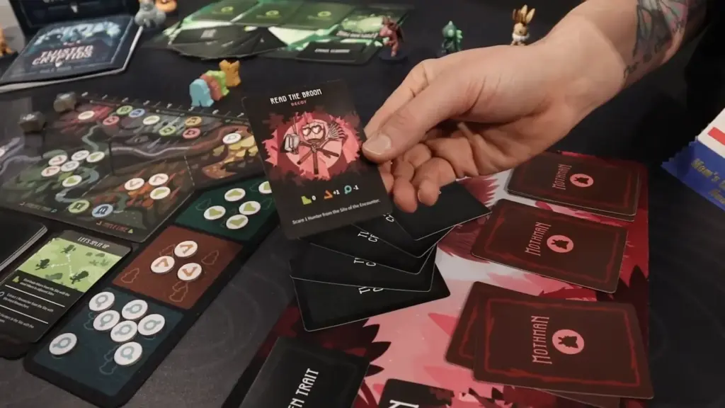 A player holding a “Read the Broom” decoy card during a demo of Twisted Cryptids at the Toy Fair NY 2025 Unstable Games showcase, showing detailed card art and tabletop components.