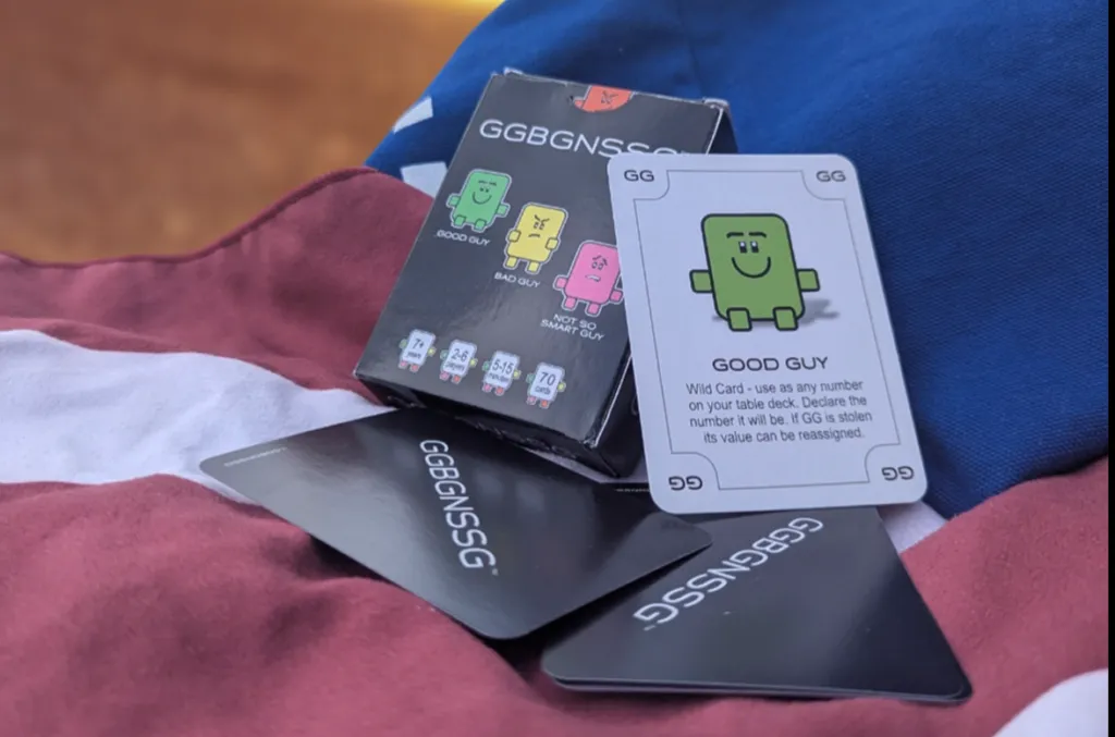 Close-up photo of the GGBGNSSG card game showing the box, several black-backed cards, and the Good Guy wild card displayed on top.