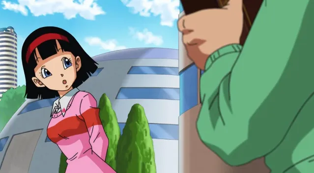 Videl standing outdoors in a pink outfit, looking surprised and curious as she glances toward Gohan, who is partially visible holding a book, with modern buildings and trees in the background.