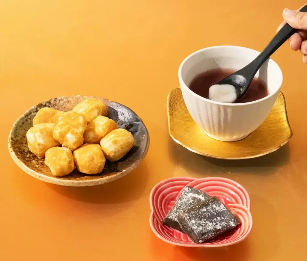 Black sesame mochi paired with a warm cup of sweet red bean soup, featuring golden mochi bites on a ceramic plate and a hand stirring mochi in tea on a gold saucer.