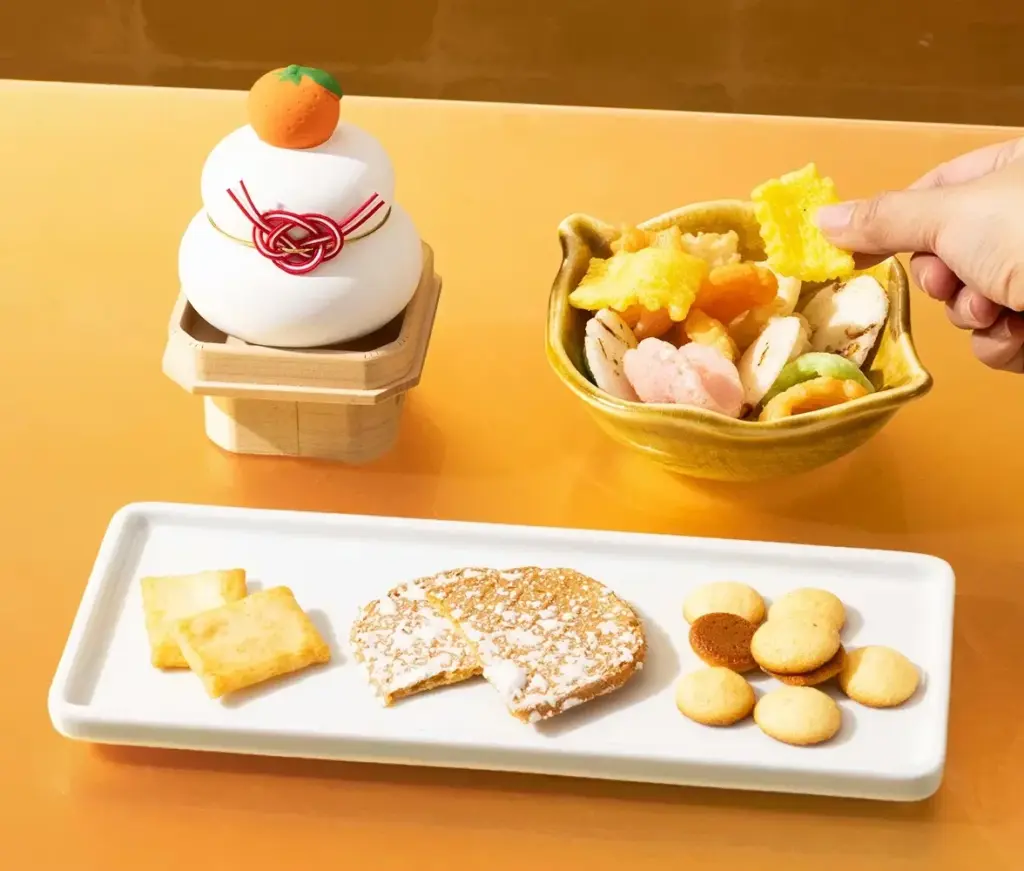 Japanese New Year snack spread featuring kagami mochi with a mandarin on top, a bowl of colorful arare rice crackers being picked up by hand, and a plate of okaki and kawara senbei on a golden table setting.