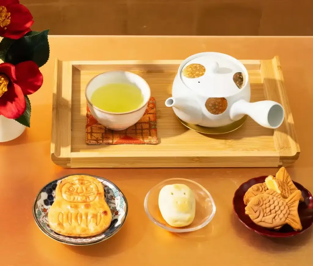A traditional Japanese tea setup with Shizuoka genmaicha served in a teacup and teapot on a wooden tray, surrounded by Daruma senbei, custard taiyaki, and assorted wagashi on small decorative plates.